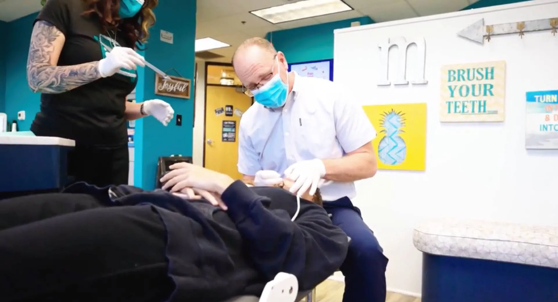 Orthodontic consultation scene at Macdonald Orthodontics, featuring a dentist in a mask examining a patient's teeth, with a dental assistant nearby, and colorful motivational signs on the wall promoting dental health.
