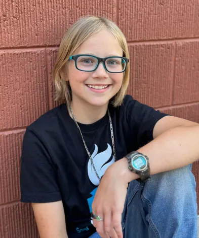 Smiling child wearing glasses and a black t-shirt, seated against a textured wall, showcasing a friendly demeanor and a casual style.