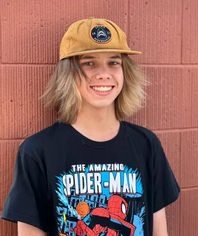 Smiling young boy wearing a brown cap and a Spider-Man t-shirt, standing against a red brick wall, representing the fun and caring atmosphere of Macdonald Orthodontics.