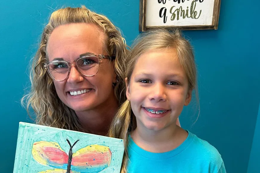 Smiling orthodontic patient with braces holding a colorful painting alongside a staff member in a dental office setting.